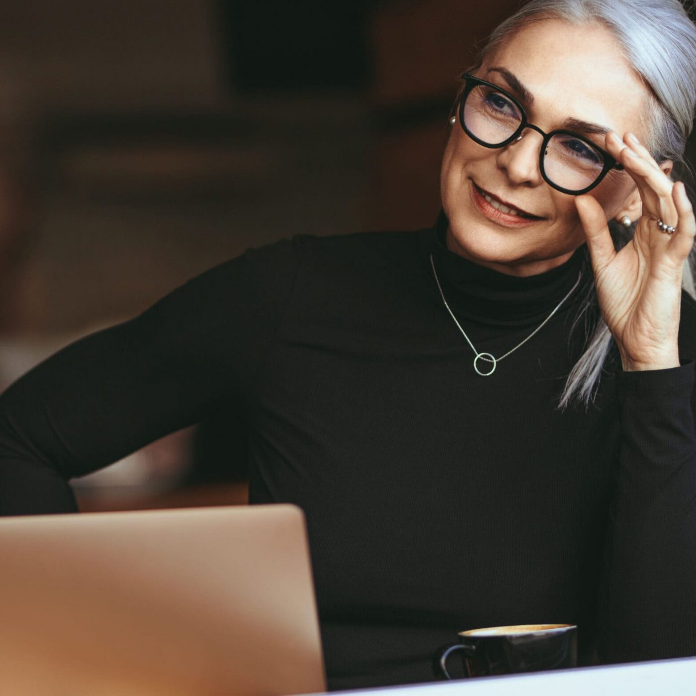 Portrait of beautiful senior businesswoman sitting at cafe with laptop looking away and thinking. Mature woman at coffee shop.